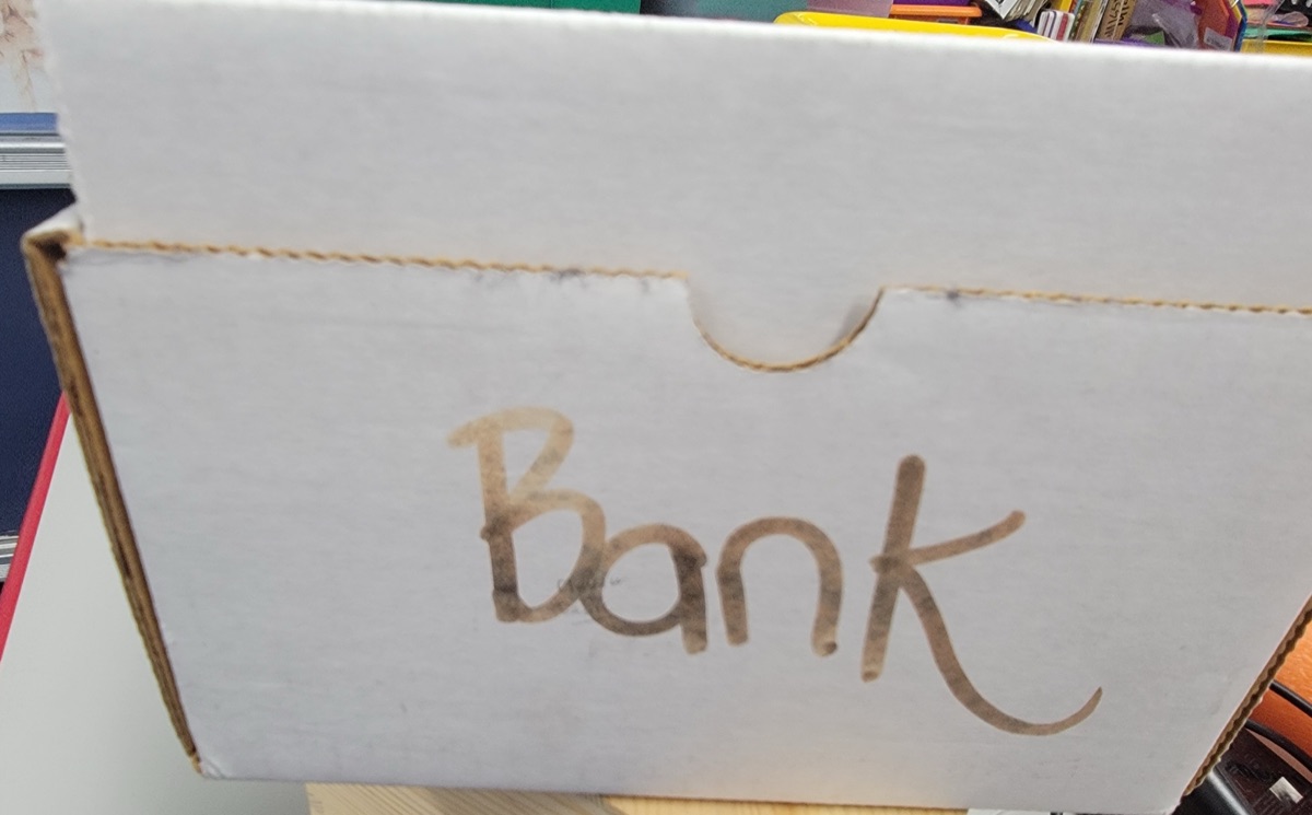 A white cardboard file box labeled Bank in marker, sitting on a shelf in Cynthia's classroom.
