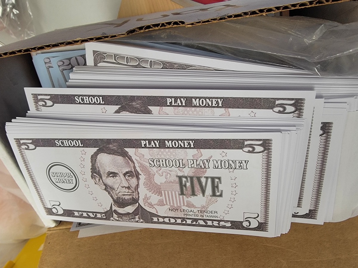 Stacks of school play money in a cardboard box, the top bills marked FIVE DOLLARS, SCHOOL PLAY MONEY and NOT LEGAL TENDER.