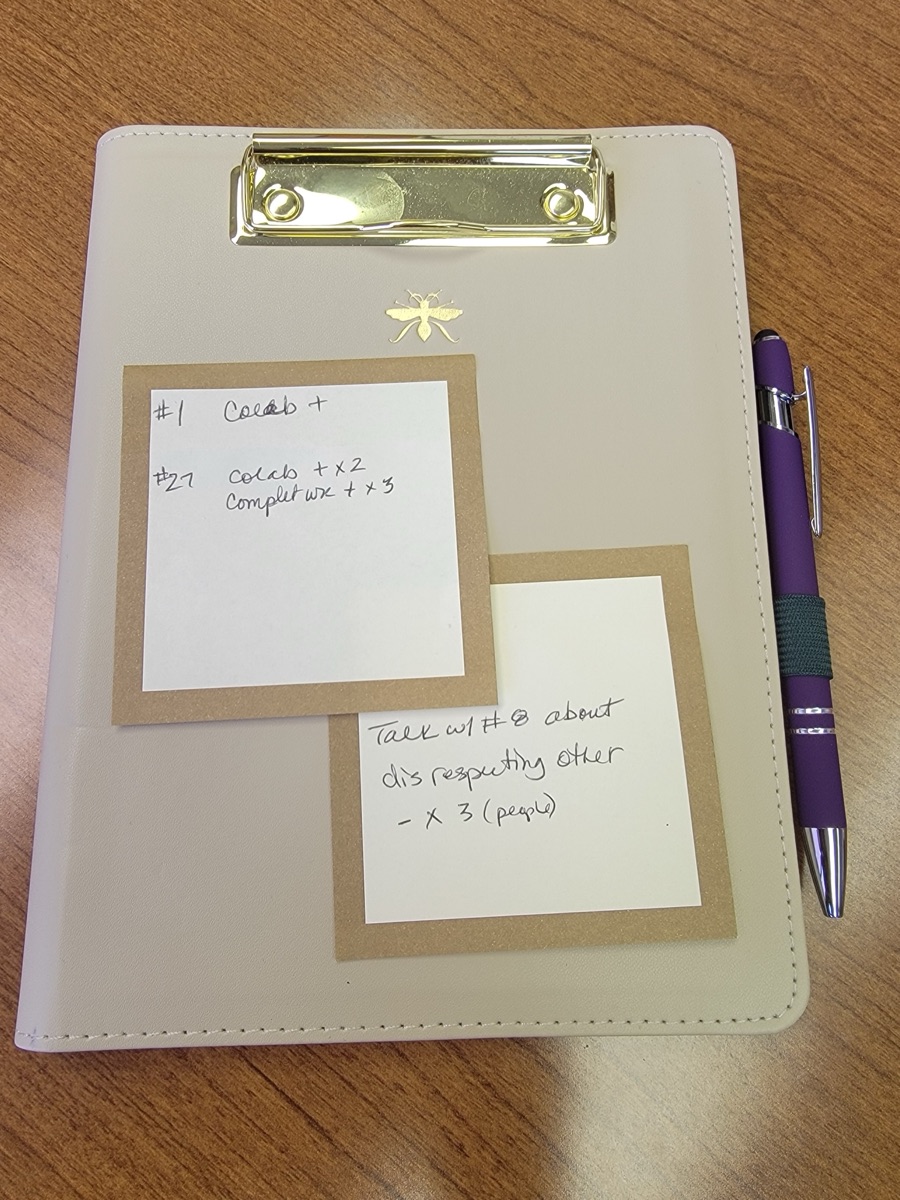 A clipboard holding two handwritten sticky notes, one recording collaboration and completed work points by student number, the other a reminder to talk with a student about disrespecting others.